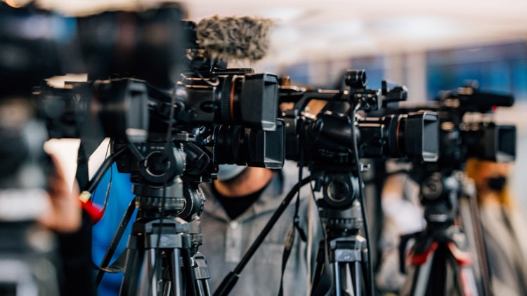 Several professional video cameras set up on tripods at a press event, with blurred people in the background.