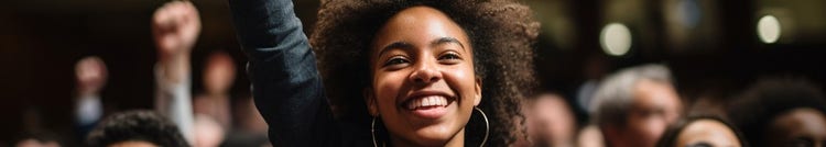 A joyful woman with curly hair raises her fist in the air during a vibrant demonstration or convention, smiling enthusiastically in a crowd of people.