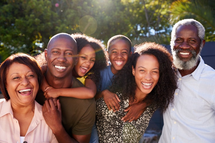 A multi-generational Black family is smiling and embracing outdoors in the bright sun.