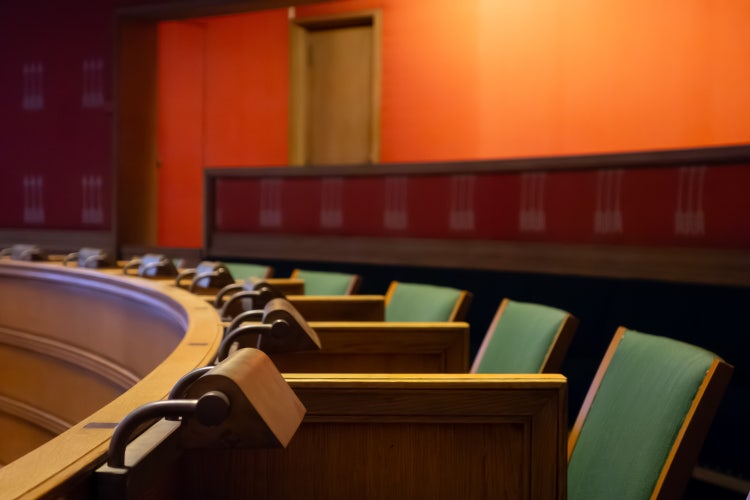 Empty curved wooden desks with built-in desk lamps and green upholstered chairs face an upper balcony area with vibrant orange walls.